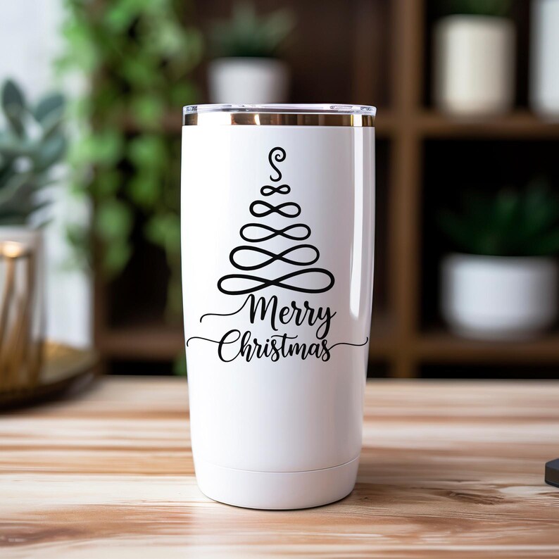 Op de afbeelding: Een witte ge&iuml;soleerde beker met een gouden rand. De beker heeft een zwart, gestileerd kerstboomontwerp en de woorden "Merry Christmas" in een scriptlettertype. De beker staat op een houten oppervlak.