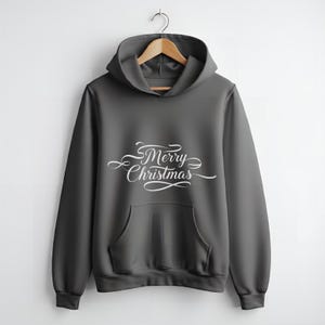 Op de afbeelding: Een donkergrijze hoodie met een voorzak en een capuchon. De woorden "Merry Christmas" staan in witte cursieve letters op de borst. De hoodie hangt aan een houten kleerhanger.