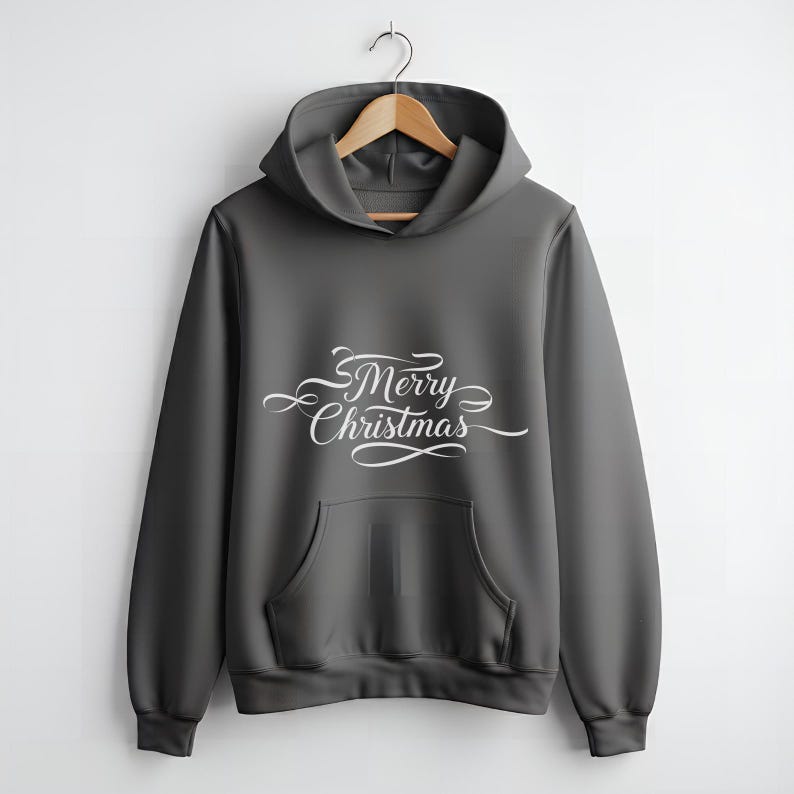 Op de afbeelding: Een donkergrijze hoodie met een voorzak en een capuchon. De woorden "Merry Christmas" staan in witte cursieve letters op de borst. De hoodie hangt aan een houten kleerhanger.