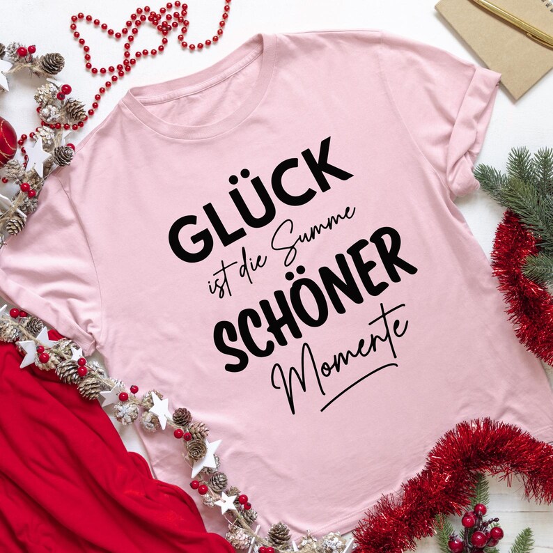 Puede incluir: Camiseta rosa claro con el texto alem&aacute;n "GL&Uuml;CK ist die Summe SCH&Ouml;NER Momente" en negro. La camiseta tiene cuello redondo y mangas cortas. Elementos decorativos: guirnaldas rojas y plateadas, pi&ntilde;as y estrellas.