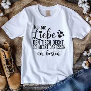 May include: White t-shirt with black text that reads "Wo die Liebe den Tisch deckt, schmeckt das Essen am besten." The text is accompanied by heart graphics. The shirt is displayed on a burlap surface.