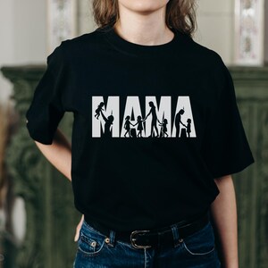 Op de afbeelding: Zwart T-shirt met het woord "MAMA" in witte blokletters. Het woord "MAMA" heeft een silhouetontwerp van een ouder en kinderen. De ouder houdt een kind omhoog en de andere kinderen houden elkaars hand vast.