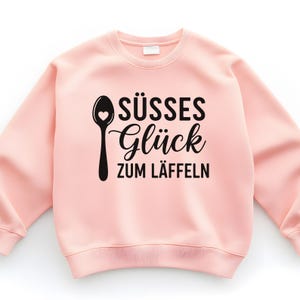 May include: A light pink sweatshirt with a black graphic of a spoon with a heart and the German text "S&Uuml;SSES Gl&uuml;ck ZUM L&Auml;FFELN". The sweatshirt has a crew neck and long sleeves.