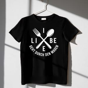 May include: Black t-shirt with a white graphic featuring a fork and spoon crossed over each other. The text "LIEBE GEHT DURCH DEN MAGEN" is printed in a circular pattern around the utensils. The shirt is hanging on a white hanger.