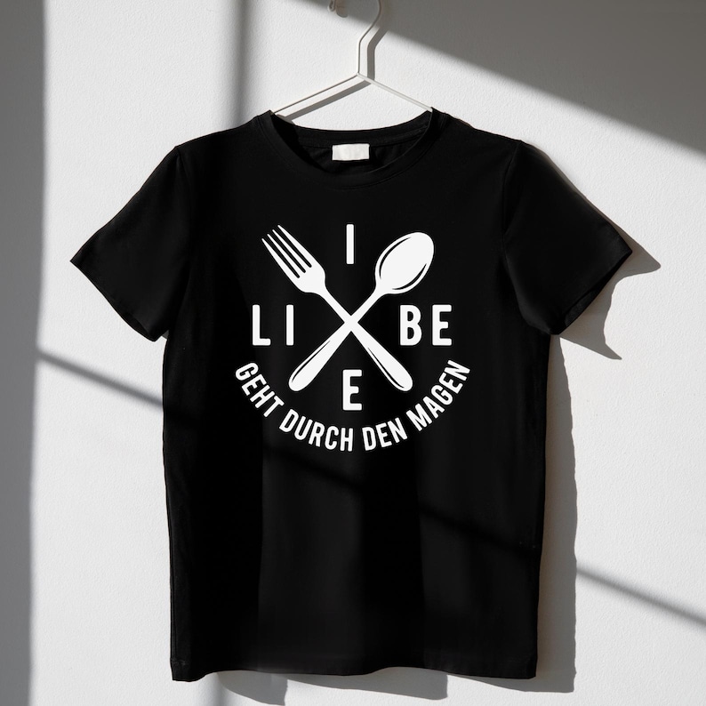 May include: Black t-shirt with a white graphic featuring a fork and spoon crossed over each other. The text "LIEBE GEHT DURCH DEN MAGEN" is printed in a circular pattern around the utensils. The shirt is hanging on a white hanger.