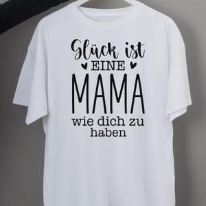 Op de afbeelding: Wit T-shirt met zwarte tekst: "Gl&uuml;ck ist eine Mama wie dich zu haben." De tekst is in een scriptletter met het woord "MAMA" in een vetgedrukte letter. Het T-shirt heeft korte mouwen.