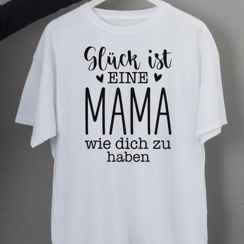 Op de afbeelding: Wit T-shirt met zwarte tekst: "Gl&uuml;ck ist eine Mama wie dich zu haben." De tekst is in een scriptletter met het woord "MAMA" in een vetgedrukte letter. Het T-shirt heeft korte mouwen.
