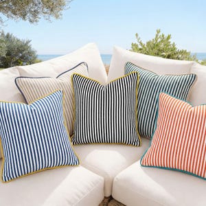 Puede incluir: Conjunto de cojines decorativos a rayas en varios colores: azul, negro, verde y naranja. Cada cojín tiene un borde en contraste. Los cojines están dispuestos sobre un sofá blanco, creando un ambiente veraniego y con estilo.