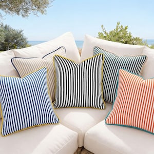 May include: A selection of striped throw cushions in various colours, including blue, black, green, and orange. The cushions have contrasting trim and are arranged on a white sofa. The background shows a view of the sea and foliage.