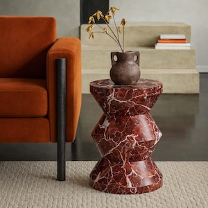 May include: A red marble side table with white veining, featuring a unique geometric design. A small, brown vase with dried flowers sits on top. The table is next to an orange sofa.
