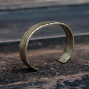 May include: A hammered brass cuff bracelet. The open-ended bracelet has a slightly textured surface and a warm, golden hue. The simple, minimalist design is suitable for everyday wear.