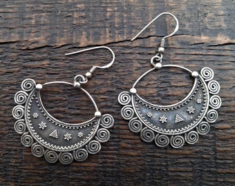 May include: A pair of silver-coloured crescent-shaped earrings. Each earring has a detailed design with spiral and dotted patterns, and small triangle shapes. The earrings have a hook closure and are displayed on a dark wooden surface.
