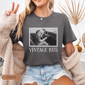 May include: A charcoal gray t-shirt featuring a rectangular black and white photograph of a young child sitting in the driver's seat of a car, holding the steering wheel. Below the image, the text VINTAGE 1975 is printed in white capital letters.