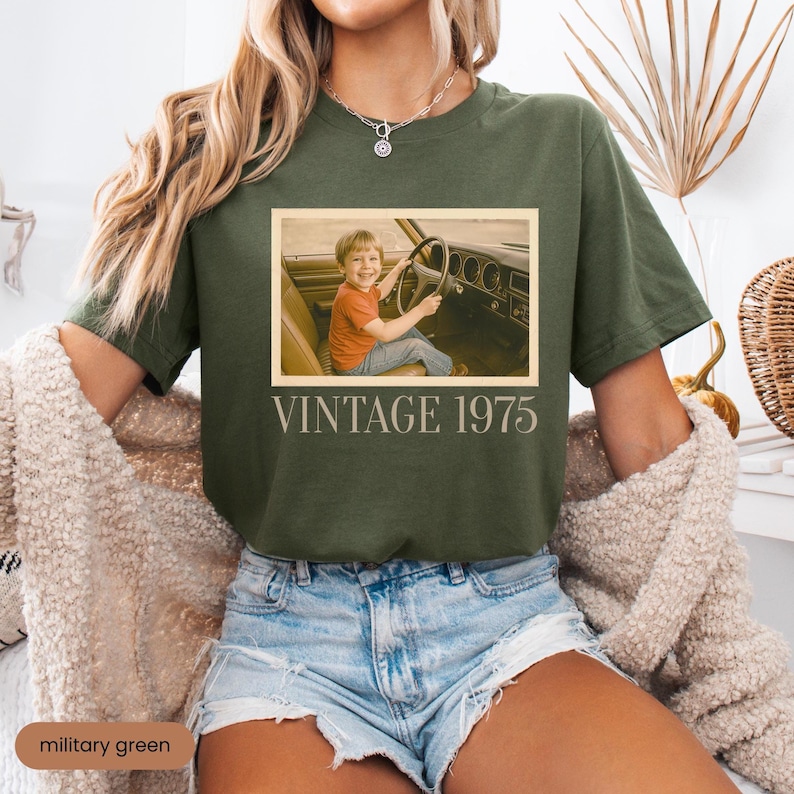 May include: A military green short-sleeve t-shirt featuring a rectangular graphic of a child sitting in the driver seat of a car. Below the image, the text VINTAGE 1975 is printed in a light beige sans-serif font. The shirt has a crew neckline.