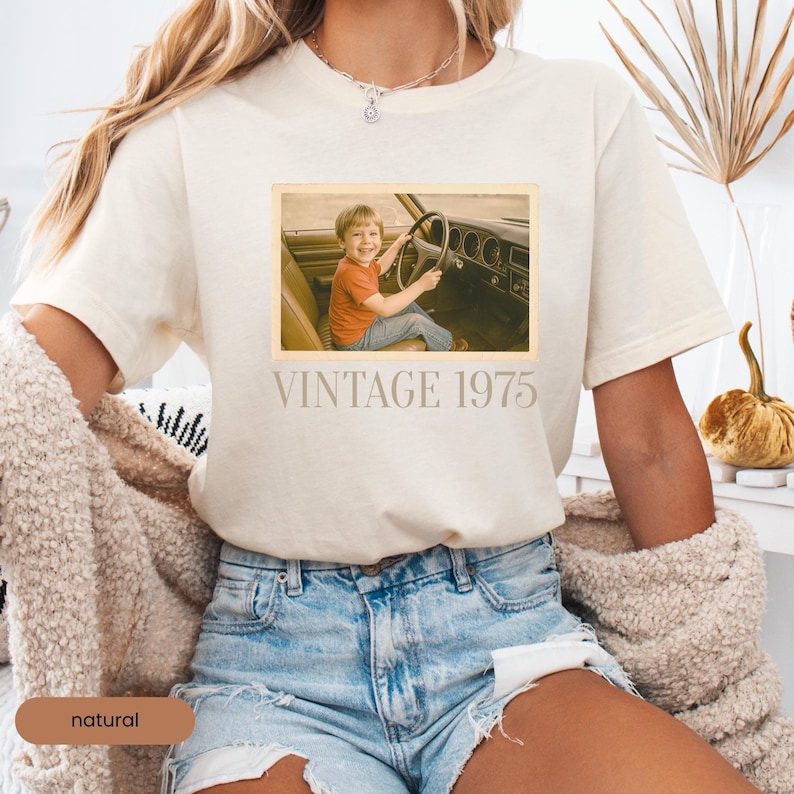 Woman wearing a natural T-shirt with a retro-style design. The shirt features a faded, vintage photo of a smiling young boy sitting in the driver’s seat of a classic 1970s car, hands on the steering wheel. Below the image is the text 'VINTAGE 1975' in a bold, serif font. The shirt has a nostalgic and personalized aesthetic