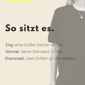 May include: A black t-shirt with a white crew neck collar worn by a person. The image features text in German: 02 / Sitz, So sitzt es, Eng: eine Gro&szlig;er kleiner w&auml;hlen, Normal: deine Standard-Gr&ouml;&szlig;e, Oversized: zwei Gr&ouml;&szlig;en gr&ouml;&szlig;er w&auml;hlen, FRIEDAFUNKEL - BERLIN.