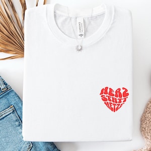 Könnte beinhalten: Weißes T-Shirt mit einem roten herzförmigen Grafik, auf dem in einer Retro-Schrift "Herz statt Hetze" steht.