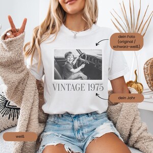 Woman wearing a white T-shirt with a retro-style design. The shirt features a faded, vintage photo of a smiling young boy sitting in the driver’s seat of a classic 1970s car, hands on the steering wheel. Below the image is the text 'VINTAGE 1975' in a bold, serif font. The shirt has a nostalgic and personalized aesthetic
