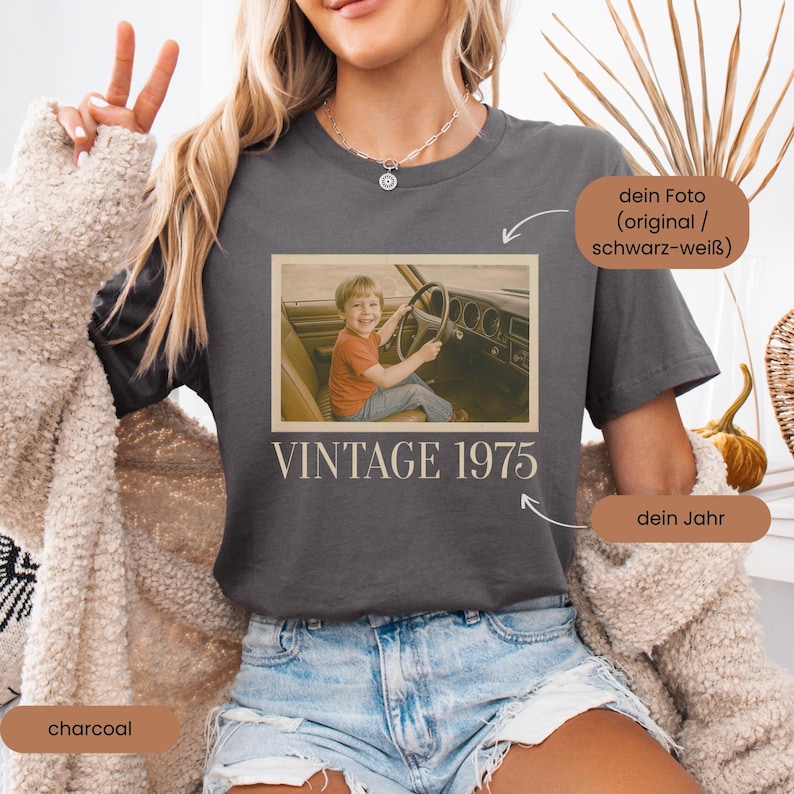 Woman wearing a dark gray T-shirt with a retro-style design. The shirt features a faded, vintage photo of a smiling young boy sitting in the driver’s seat of a classic 1970s car, hands on the steering wheel. Below the image is the text 'VINTAGE 1975' in a bold, serif font. The shirt has a nostalgic and personalized aesthetic