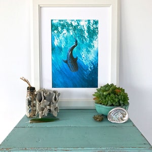 May include: A framed print of a black and white whale shark swimming in blue water with white foam. The print is in a white frame and is sitting on a light blue table with a small succulent plant in a blue pot, a seashell, and a glass jar with seashells and twine.