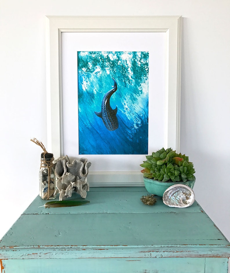 May include: A framed print of a black and white whale shark swimming in blue water with white foam. The print is in a white frame and is sitting on a light blue table with a small succulent plant in a blue pot, a seashell, and a glass jar with seashells and twine.