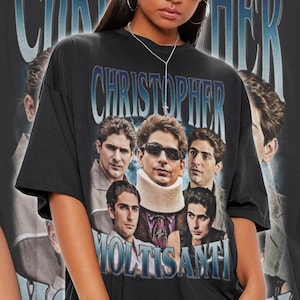 May include: Black unisex t-shirt featuring a graphic of several men with the name "CHRISTOPHER MOLTISANTI" in blue text. The shirt has a vintage style and is worn by a person wearing sunglasses and a necklace.