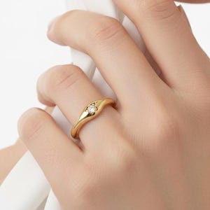 May include: A gold ring with a small, round, clear gemstone set in the center. The ring has a smooth, curved design and is worn on a finger. The ring is a piece of jewelry.