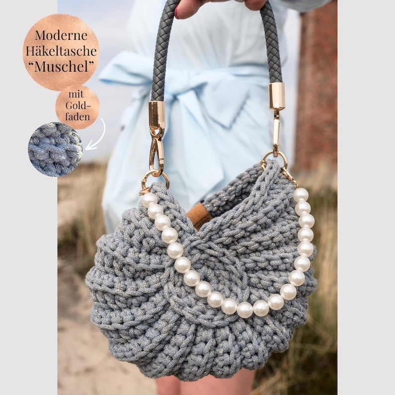 May include: A grey crocheted handbag with a shell-like shape, featuring a braided grey handle with gold-tone hardware and a decorative strand of white faux pearls draped across the front. The bag is textured with a visible yarn stitch pattern.
