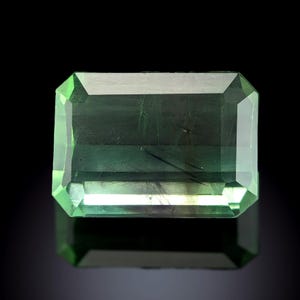 Puede incluir: Una gema rectangular, talla esmeralda, con un tono verde vibrante. La gema presenta un diseño facetado, mostrando su claridad y brillo. La piedra se encuentra sobre un fondo oscuro, resaltando su color y forma.