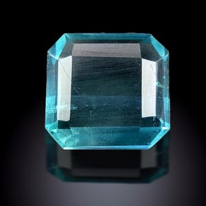 Puede incluir: Una gema rectangular facetada de color verde azulado claro. La gema tiene una superficie reflectante y está sobre un fondo negro. El corte de la gema es un corte esmeralda, con una serie de facetas planas.