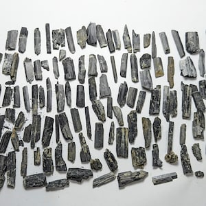 May include: A collection of raw, black tourmaline crystals of varying sizes and shapes, scattered on a white surface. The crystals have a rough, textured appearance, with shades of gray and black.