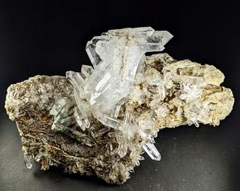 Può includere: Un gruppo di cristalli di quarzo trasparente, con terminazioni appuntite, è posto su una matrice ruvida beige e marrone. I cristalli sono traslucidi, catturando la luce e creando un effetto scintillante. È un esemplare geologico naturale.