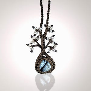 May include: A handcrafted macrame necklace featuring a tree design. The brown macrame tree has clear crystal accents and a blue and white stone pendant. The necklace hangs from a brown cord.