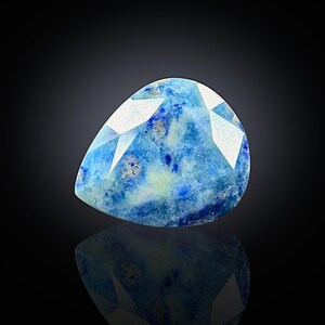 May include: A pear-shaped gemstone with a faceted top. The stone features a mix of blue hues with white and gray inclusions. The gem is set against a dark background, with a reflection visible below.