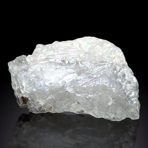 Può includere: Un esemplare minerale traslucido di forma irregolare con una struttura cristallina. La pietra è prevalentemente trasparente con sottili lampi iridescenti, su uno sfondo scuro. La superficie ha un aspetto ruvido e strutturato.