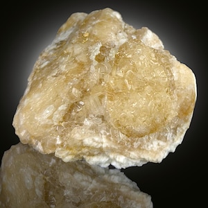 Pode incluir: Uma aproximação de um espécime mineral cristalino bruto. A pedra é de cor amarelo pálido com áreas translúcidas e uma superfície refletora por baixo. O mineral tem uma superfície texturizada e irregular com uma forma natural e orgânica.