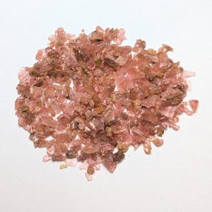 May include: A pile of small, translucent pink crystals with some brown and tan inclusions. The crystals are irregularly shaped and appear to be raw or unpolished. The crystals are scattered on a white surface.