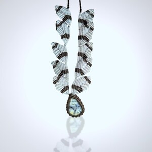May include: A handmade macrame necklace featuring a teardrop-shaped pendant with a light blue and white stone. The necklace has a unique design with white and brown woven elements, creating a feather-like pattern. The pendant is encased in a brown macrame setting.