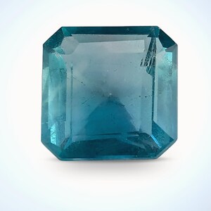 Puede incluir: Una gema rectangular facetada de color azul verdoso. La gema tiene un diseño de talla escalonada con un borde ligeramente biselado. La piedra es transparente, con inclusiones internas visibles y una superficie pulida. Sobre un fondo blanco.
