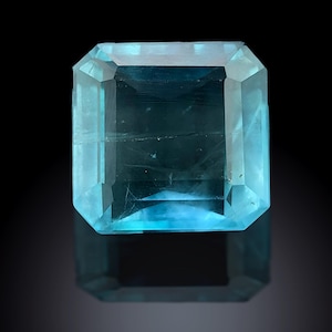 Puede incluir: Una gran gema de corte rectangular con un tono azul claro. La gema está sobre un fondo negro, con un reflejo visible debajo. Las facetas de la piedra están claramente definidas, captando la luz y creando un efecto brillante.