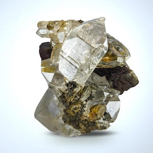 May include: A cluster of clear quartz crystals with sharp, geometric facets. The crystals are partially embedded in a matrix of dark brown and green rock. The overall appearance is translucent with light reflecting off the crystal surfaces.