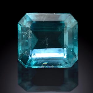 Puede incluir: Una gema de color turquesa, de corte rectangular y con superficie facetada. La gema está sobre un fondo negro, con un reflejo visible debajo. Las facetas de la piedra captan la luz, creando un efecto brillante. La forma general es un octágono.