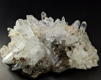 Può includere: Un gruppo di cristalli di quarzo trasparente con terminazioni appuntite e affilate. I cristalli sono disposti su una matrice ruvida, grigio-marrone, creando un aspetto strutturato. Il quarzo riflette la luce, mostrando la sua trasparenza e formazione naturale. Lo sfondo è nero.