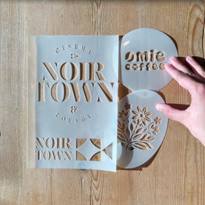 May include: A selection of stencils on a wooden surface. The stencils feature the text "NOIR TOWN & LOUNGE" and "Omie coffee", alongside a floral design. The stencils are made of a translucent material.