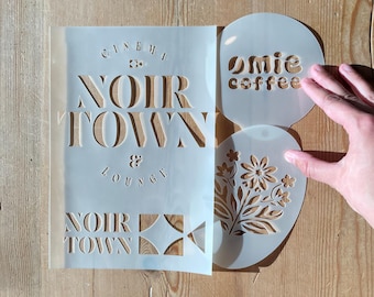 May include: A collection of stencils on a wooden surface. The stencils include text that reads "NOIR TOWN & LOUNGE" and "Omie coffee", along with a floral design. The stencils are made of a translucent material.