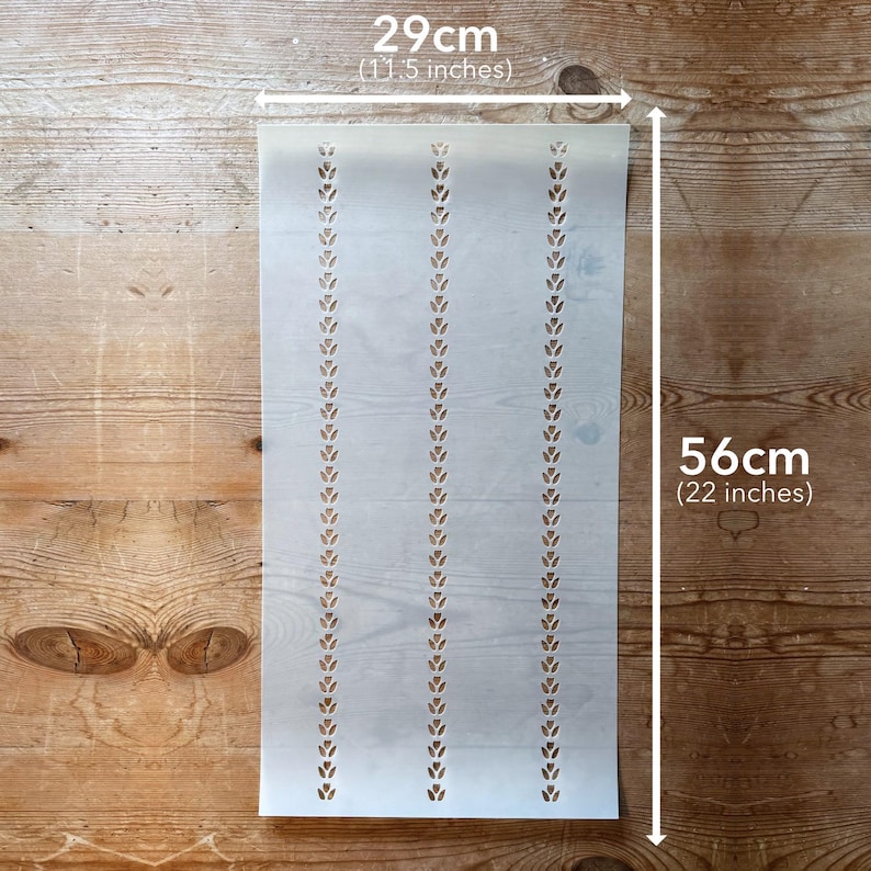 May include: A stencil with a repeating leaf pattern, 11.5 inches wide and 22 inches long. The stencil is light in color and has three vertical rows of leaf-shaped cutouts. The stencil is placed on a wooden surface.