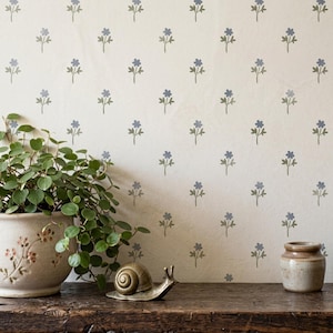 Op de afbeelding: Een crèmekleurige muur met een herhalend patroon van kleine, gedempte blauwe bloemen met groene stelen. Een keramische pot met een groene plant, een metalen slakfiguurtje en een kleine bruine keramische pot staan op een donkerhouten oppervlak.