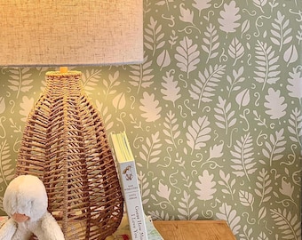 May include: A woven wicker lamp with a beige lampshade sits on a wooden surface next to a stack of books and a stuffed animal. The background features a green wallpaper with a white leaf pattern. One book is titled "Guess How Much I Love You."