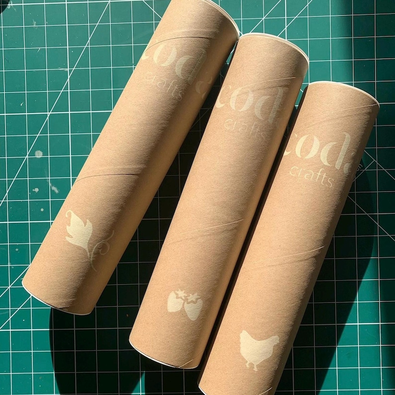 May include: Three cylindrical cardboard tubes with the text "cod crafts" printed on them. Each tube has a different white graphic: a leaf, strawberries, and a chicken. The tubes are arranged on a green cutting mat.
