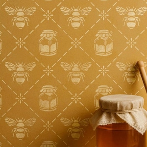 Può includere: Muro color senape con un motivo ripetuto di api, barattoli di miele e fiori. Un barattolo di miele con una copertura di stoffa e un cucchiaio di miele di legno sono in primo piano. Il motivo è in una tonalità di giallo più chiara.